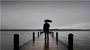 Ein Mann steht mit Regenschirm auf einem Steg. Man sieht nur seine Silhouette. Ringsherum ist Wasser. Der Himmel ist grau und wolkenverhangen.