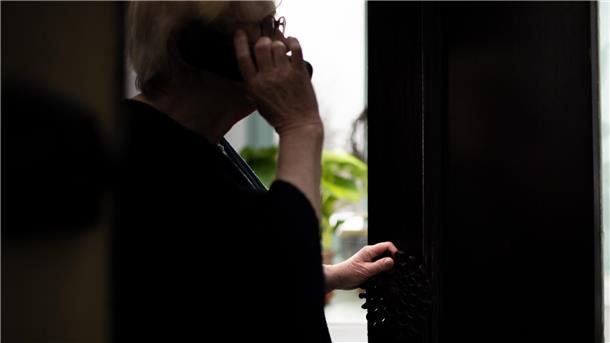 Eine Seniorin hält ein Telefon am Ohr und öffnet ihre Haustür einen Spalt weit.