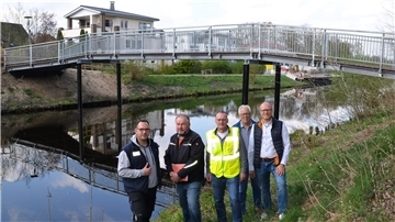 Fünf Männer stehen vor einer neu errichteten Fußgängerbrücke über den Hadelner Kanal in Geestland. Hinter ihnen verläuft die Brücke mit Metallgeländer über das Wasser. 