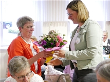 Eine Frau in oranger Strickjacke bekommt einen Blumenstrauß überreicht.