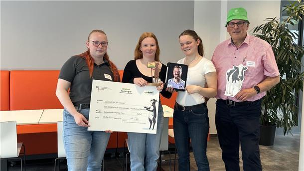 Beerster Auszubildende überzeugen mit Projekt zur Biolandwirtschaft Drei Schülerinnen des GenoKollegs Münster stehen mit Lehrer Klaus Feils in einem modernen Raum. Sie halten einen Scheck über 750 Euro, einen Pokal, ein Tablet mit einem Porträt und eine Kuhfigur – als Auszeichnung für den dritten Platz beim Wettbewerb „Echt kuh-l! 2025“.