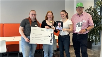 Drei Schülerinnen des GenoKollegs Münster stehen mit Lehrer Klaus Feils in einem modernen Raum. Sie halten einen Scheck über 750 Euro, einen Pokal, ein Tablet mit einem Porträt und eine Kuhfigur – als Auszeichnung für den dritten Platz beim Wettbewerb „Echt kuh-l! 2025“.