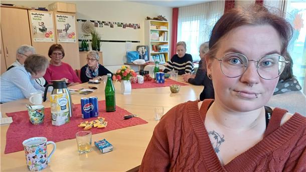 Eine jüngere Frau blickt in die Kamera, hinter ihr diskutieren ältere Frauen an einem großen Tisch. 