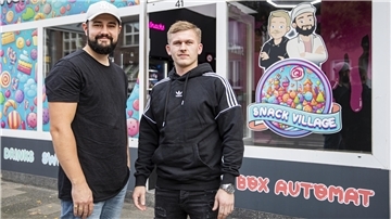 Ein Mann mit dunklem Bart und Basecap und ein Mann mit blonden Haaren stehen vor einem Kiosk namens Snack Village.