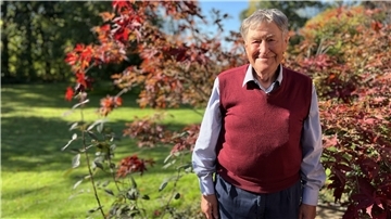Ein älterer Herr mit rotem Pollunder und Hemd steht vor rotem Ahorn. Im Hintergrund sein Garten.