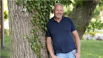 Ein Mann lehnt an einem Baum und schaut in die Kamera.