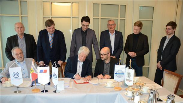Viele Männer sitzen und stehen um einen Tisch und unterzeichnen eine Absichtserklärung.