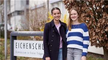Zwei Schülerinnen stehen vor dem Eingangsschild ihrer Schule und lächeln in die Kamera.