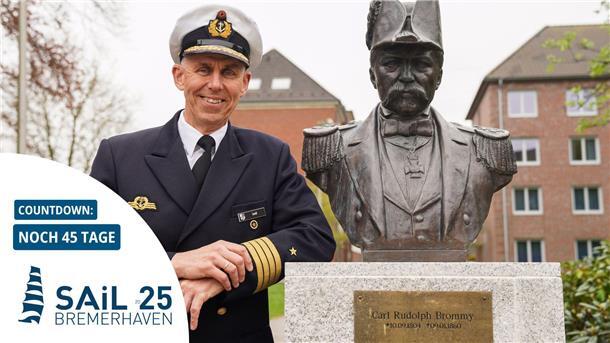 Ein Marineoffizier in Uniform steht lächelnd neben einer Büste von Carl Rudolph Brommy; im Vordergrund ein Countdown zur Sail 2025 in Bremerhaven mit „Noch 45 Tage“.