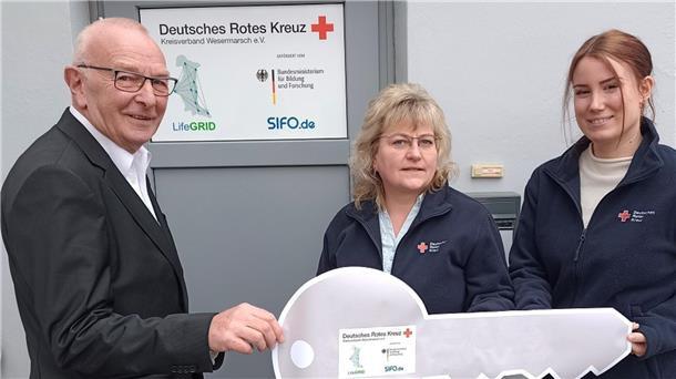Karl-Heinz Röben, Vorsitzender des DRK-Kreisverbandes Wesermarsch, übergibt Kathleen Hirsch und Chariece Atli als Expertin für die Pflege symbolisch den Schlüssel für die neuen Geschäftsräume des DRK-LifeGRID-Teams in der Breiten Straße 104 in Brake.