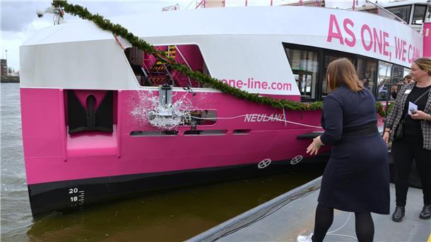 Eine Frau lässt eine Sektflasche am Bug einer pink-weiß gestrichenen Fähre mit dem Namen "Neuland" zerschellen.