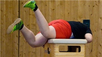 Ein übergewichtiger Junge rollt sich im Sportunterricht über einen Kasten. 