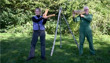 Eine Frau und ein Mann auf einer Wiese mit Büschen im Hintergrund. Beide halten verschiedene Hölzer in den Händen.