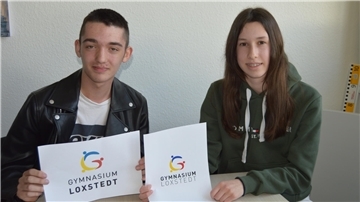 Schüler und Schülerin halten das auf Papier gedruckte neue Logo des Gymnasiums Loxstedt hoch