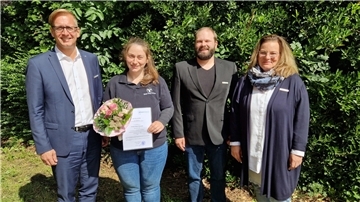 Vier Menschen stehen vor einer grünen Hecke. Landrat Stephan Siefken beglückwünscht Wiebke Mayer-Reinecke zu ihrer neuen Aufgabe als Bezirksschornsteinfegermeisterin.