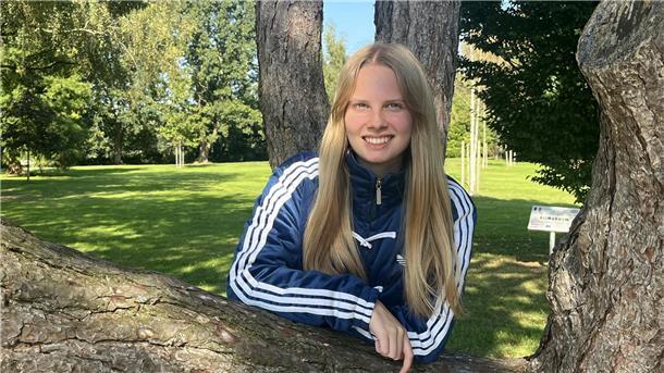 Eine junge Frau lehnt an einem Baum und lächelt in die Kamera.