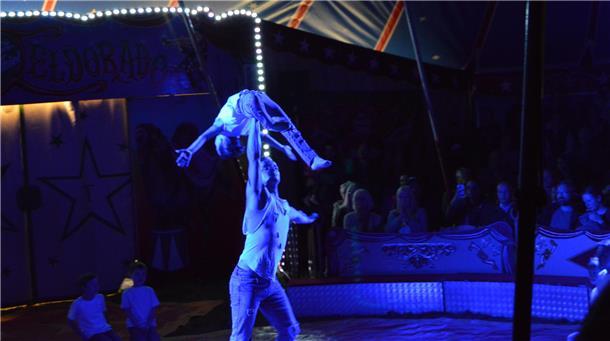 Ein Schüler balanciert beim Zirkusprojekt in Stotel auf der in die Höhe gestreckten Hand eines Akrobaten. Die Szene in der Manege ist in blaues Licht getaucht.