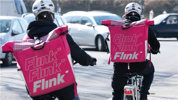 Zwei Fahrradkuriere tragen Rucksäcke mit dem Logo des Lebensmittel Lieferdienstes Flink.