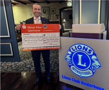 Zum Jubiläum: Lions Bederkesa fördern Projekte mit 25.000 Euro Ein Mann im Anzug hält einen großen symbolischen Spendenscheck über 25.000 Euro in den Händen. Neben ihm steht ein beleuchtetes Schild mit dem Logo des Lions Club Bederkesa.
