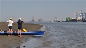 Bild von Ruderern mit ihrem Boot auf einer Sandbank in der Weser. Im Hintergrund sind Großsegler und die Hafenkräne in Bremerhaven zu sehen.