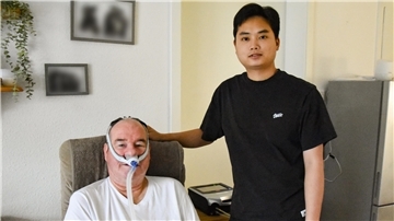 Ein älterer Mann mit Maskenbeatmung sitzt lächelnd auf einem Sessel, neben ihm steht ein junger Mann.