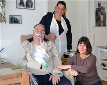 Ein Mann mit Beatmungsmaske sitzt mit wachem Blick in einem Sessel, hinter und neben ihm sind zwei Frauen, im Hintergrund ist das Beatmungsgerät erkennbar.