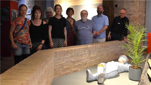 Gruppe von neun Personen steht in einem Ausstellungsraum hinter einer niedrigen Backsteinmauer. Im Vordergrund liegt eine Figur in Mosaikoptik neben einer Topfpflanze.