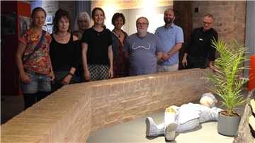 Gruppe von neun Personen steht in einem Ausstellungsraum hinter einer niedrigen Backsteinmauer. Im Vordergrund liegt eine Figur in Mosaikoptik neben einer Topfpflanze.