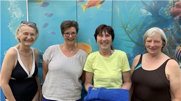 Vier Frauen stehen lächelnd vor einer Wand mit Unterwasser-Motiven. Eine von ihnen hält ein blaues Handtuch in den Händen. Es handelt sich um die Begrüßung des 400. Mitglieds im Förderverein Freibad Grünhöfe.