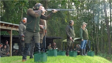 Mehrere Jäger stehen auf einem Schießstand im Grünen, tragen Gehörschutz und Jagdbekleidung. Einer von ihnen zielt mit einer Flinte auf eine fliegende Wurfscheibe, während andere warten. Im Hintergrund beobachten weitere Personen das Geschehen.