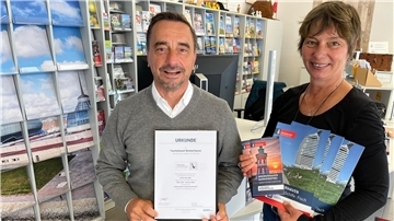 Ein Mann im grauen Pullover und eine Frau in schwarzem Shirt stehen in einem Tourismusbüro. Der Mann hält eine Urkunde mit dem Titel „Tourismusort Bremerhaven“, die Frau zeigt mehrere barrierefreie Informationsbroschüren über Bremerhaven.