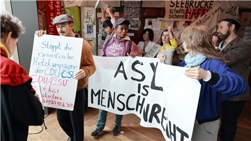 Demonstrationsszene mit Protestplakaten in einer Ausstellung; mehrere Figuren halten Schilder mit Aufschriften wie „ASYL IST MENSCHENRECHT“ und „Stoppt die rassistische Hetzkampagne der CDU/CSU“.
