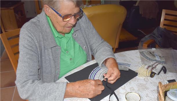 Eine alte Frau sitzt an einem Tisch und hat Kragen aus Perlen vor sich liegen.