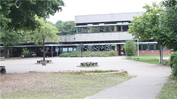 Blick auf den Schulhof und das Gebäude der Grundschule Beverstedt mit Bäumen im Vordergrund. An den Fenstern hängen bunte Girlanden und Papierdekorationen.