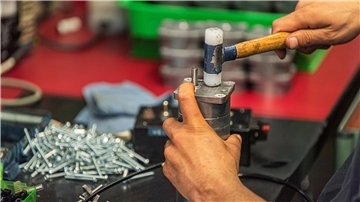 Eine Hand, die mit einem Hammer auf ein Metallteil schlägt. Die andere Hand hält das Metallteil fest. Im Hintergrund liegen Schrauben.
