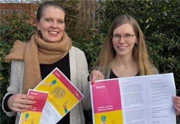 Monika van Büren und Judith Bremer stehen vor einer grünen Hecke und halten Papiere des "klimafit"-Kursus hoch