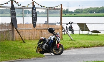 Ein Motorrad steht vor einer Grünfäche. Im Hintergrund ist zusammengebundenes Schilf zu erkennen, das ein Strandhaus formt. Auch eine Wasserfläche ist sichtbar.