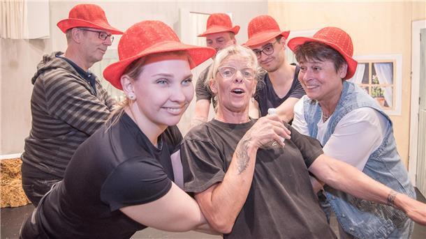 Eine Gruppe von sechs Menschen, teils mit roten Hüten, steht eng zusammen und posiert lachend für die Kamera. Im Vordergrund werden eine Frau in Schwarz und eine Frau in Jeansweste von den anderen gestützt.