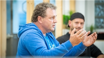 Ein Mann in blauem Pulli gestikuliert, im Hintergrund ist ein Mann mit schwarzem Pulli und Kappe zu sehen.