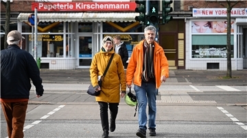 Eine Frau mit Brille und gelber Jacke sowie ein Mann mit Brille und orangefarbener Jacke. Er trägt in seiner rechten Hand einen Helm. Beide gehen über eine Straße, die Ampel zeigt grünes Licht.