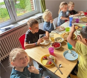 Mehrere Kinder sitzen fröhlich an einem Tisch und essen frisch gebackene Waffeln mit Sahne, Erdbeeren und Bananen. Auf dem Tisch stehen bunte Becher und Teller sowie Schüsseln mit Obst.