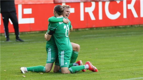 Zwei Männer knien auf dem Fußballrasen und liegen sich lachend in den Armen.
