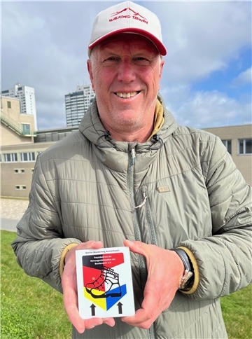 Ein Mann mit Mütze lächelt und hält zeigt mit beiden Händen ein handgroßes Schild. Neue Beschilderung für Bremerhavens einzigen offiziellen Wanderweg: Frank Reininghaus wandert mit einer Aluleiter 125 Kilometer durch die Stadt, um 100 Tafeln anzubringen