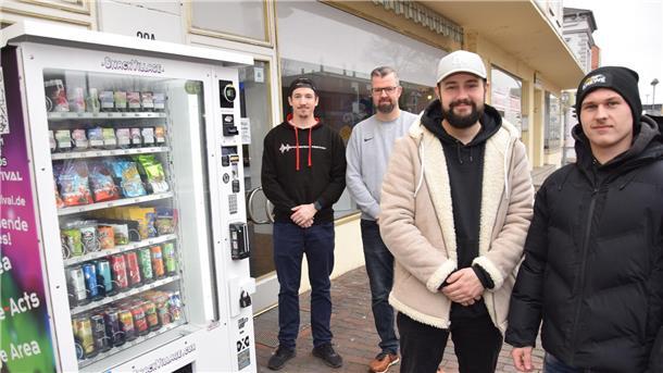 Vier Männer stehen neben einem Automaten mit Getränken und Snacks.
