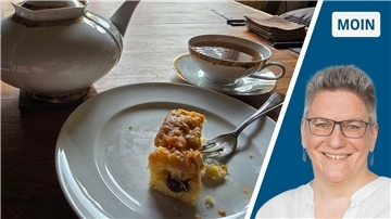Ein Stück Kuchen liegt auf einem Teller auf einem Wohnzimmertisch. Daneben steht eine Teekanne und eine volle Teetasse.