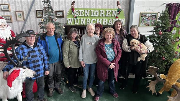 Sechs Frauen, zwei Männer und ein Hund stehen in einer weihnachtlich eingerichteten Ecke einer Halle und lächeln in die Kamera.