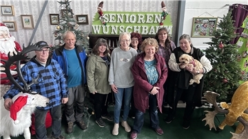 Sechs Frauen, zwei Männer und ein Hund stehen in einer weihnachtlich eingerichteten Ecke einer Halle und lächeln in die Kamera.