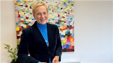 Blonde Frau mit Blazer vor buntem Gemälde lächelt in die Kamera.