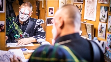 Ein geschminkter Clown sitzt vor einem Spiegel und blickt auf einen Zettel auf einem Tisch.