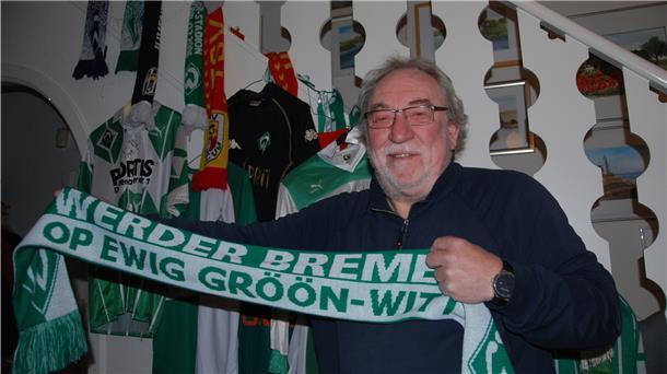Ein Mann steht vor einem Treppenaufgang mit lauter Fanutensilien. In der Hand hält er einen Schal des SV Werder Bremen.